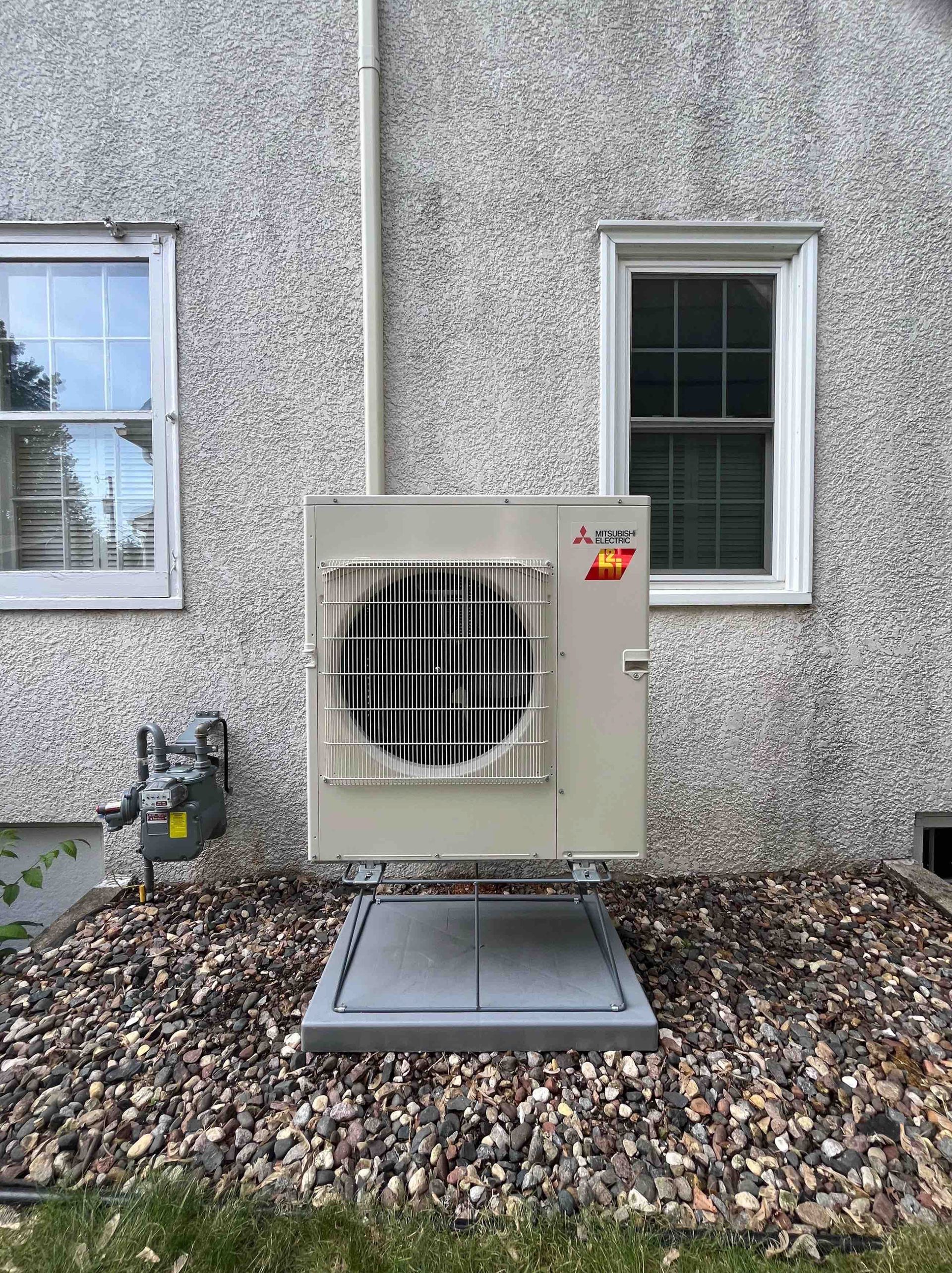 An outdoor HVAC unit on a gravel base, mounted on a stucco wall, with two windows on either side.