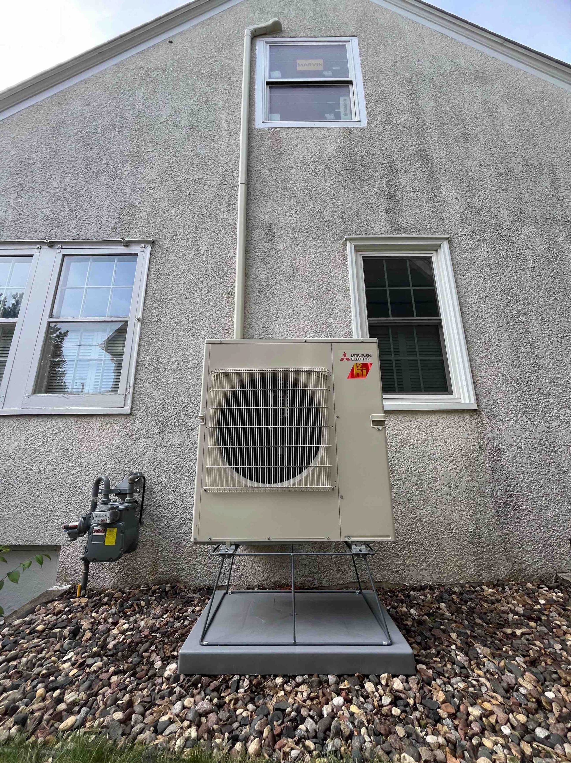 An outdoor air conditioning unit mounted on a platform, with a gas meter to the left, and windows above.