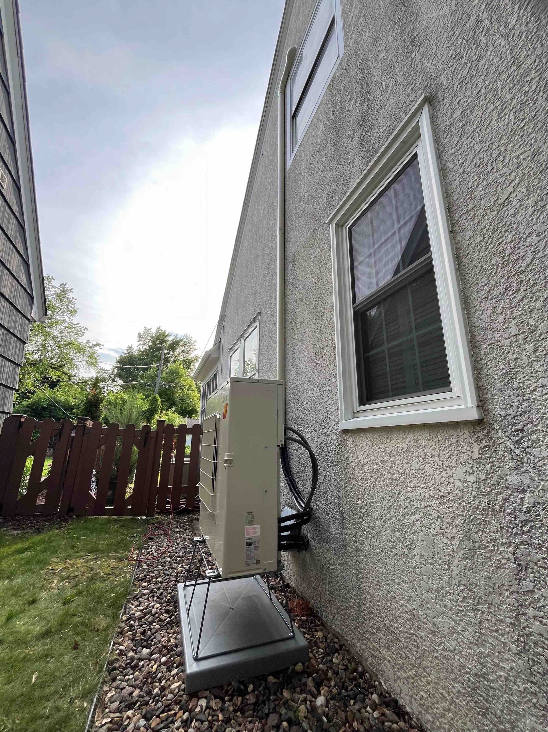 An air conditioning unit mounted on a house exterior next to a window and brown fence.