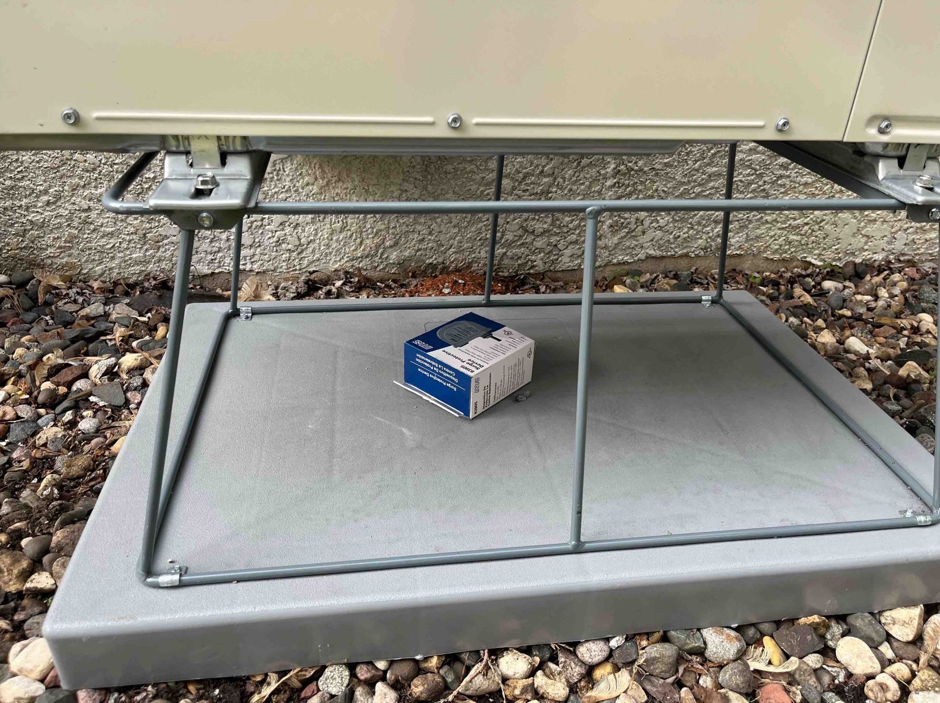 Blue electrical box on a gray pad under a beige structure, protected by a metal cage, outside.