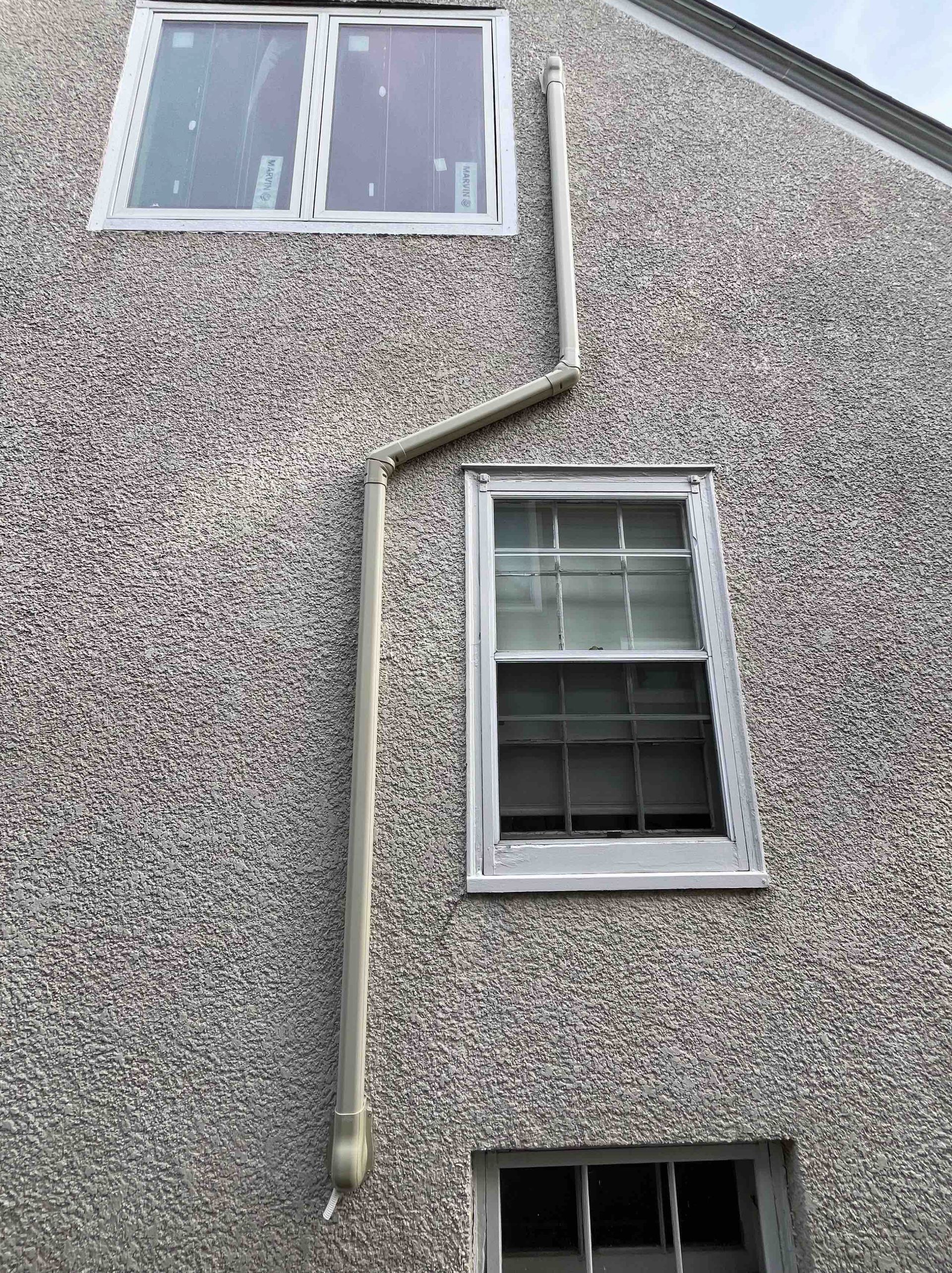 Light-colored rain gutter runs down the side of a house with three windows. The siding has a rough texture.