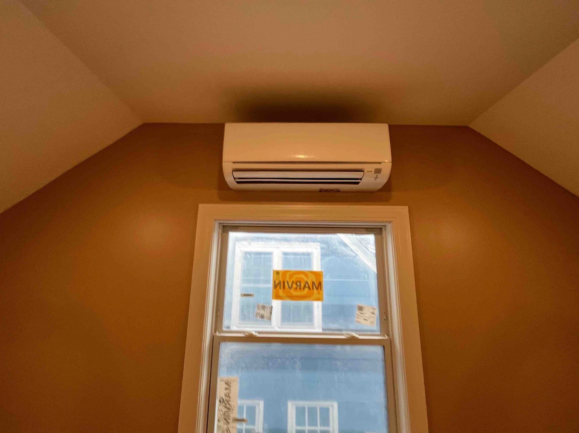 An air conditioning unit installed above a window in a room with brown walls.