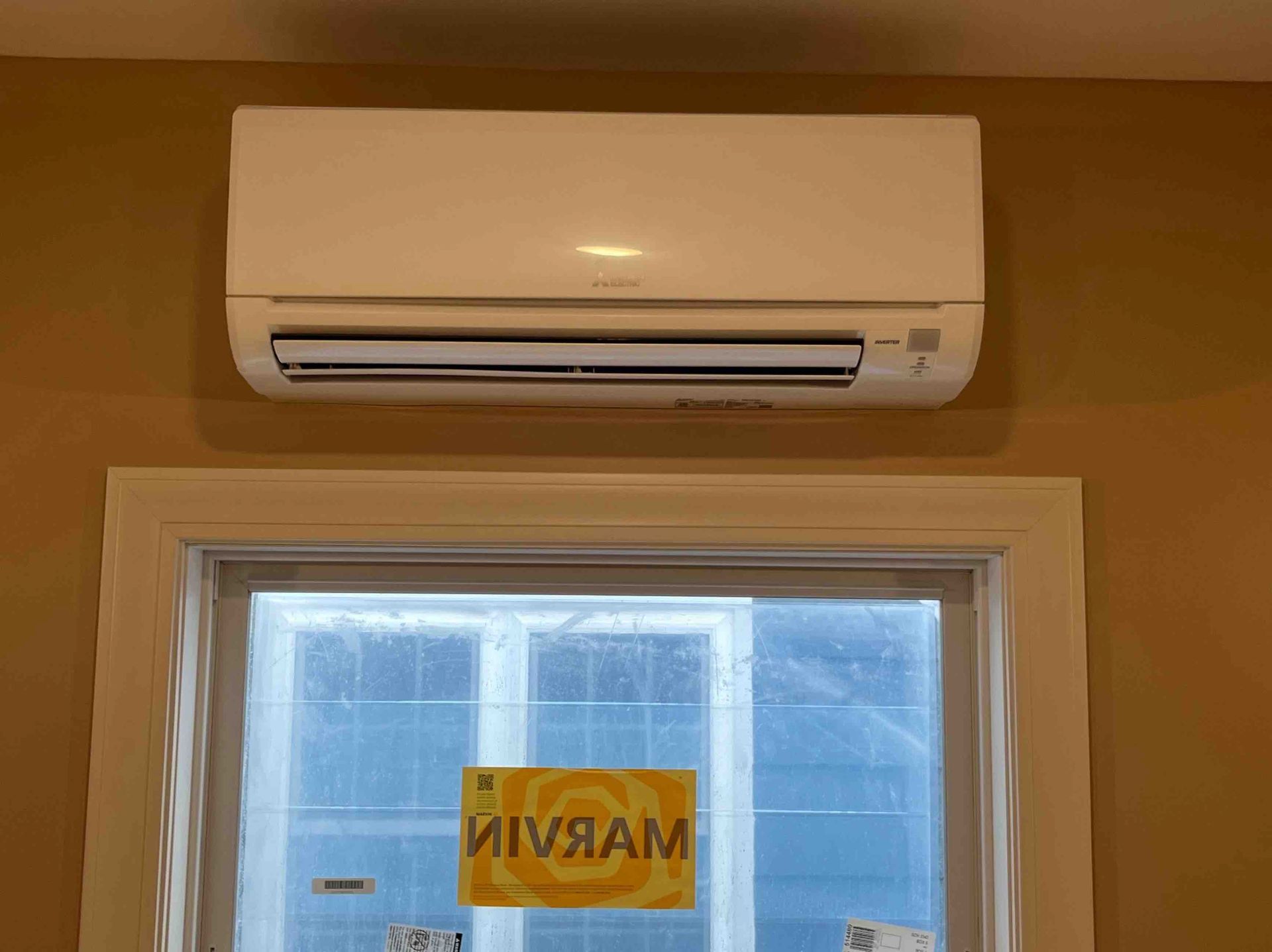 White ductless air conditioner mounted above a window on a tan wall.