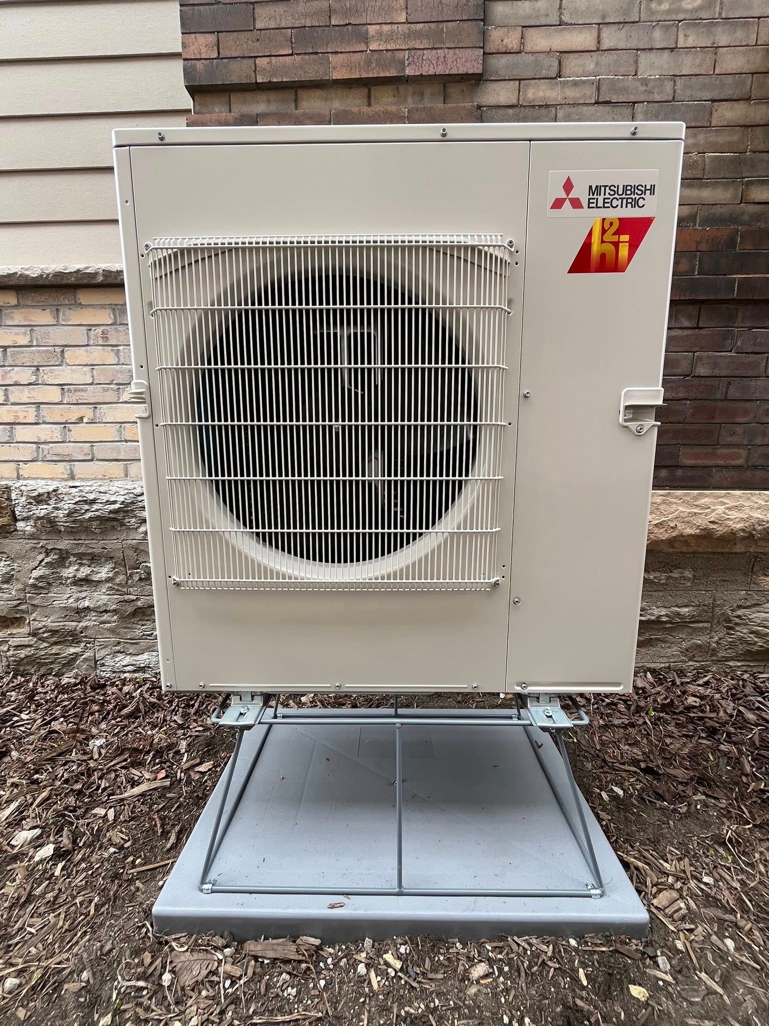 Mitsubishi heat pump unit, beige with metal grille, mounted on a gray base against a brick wall.