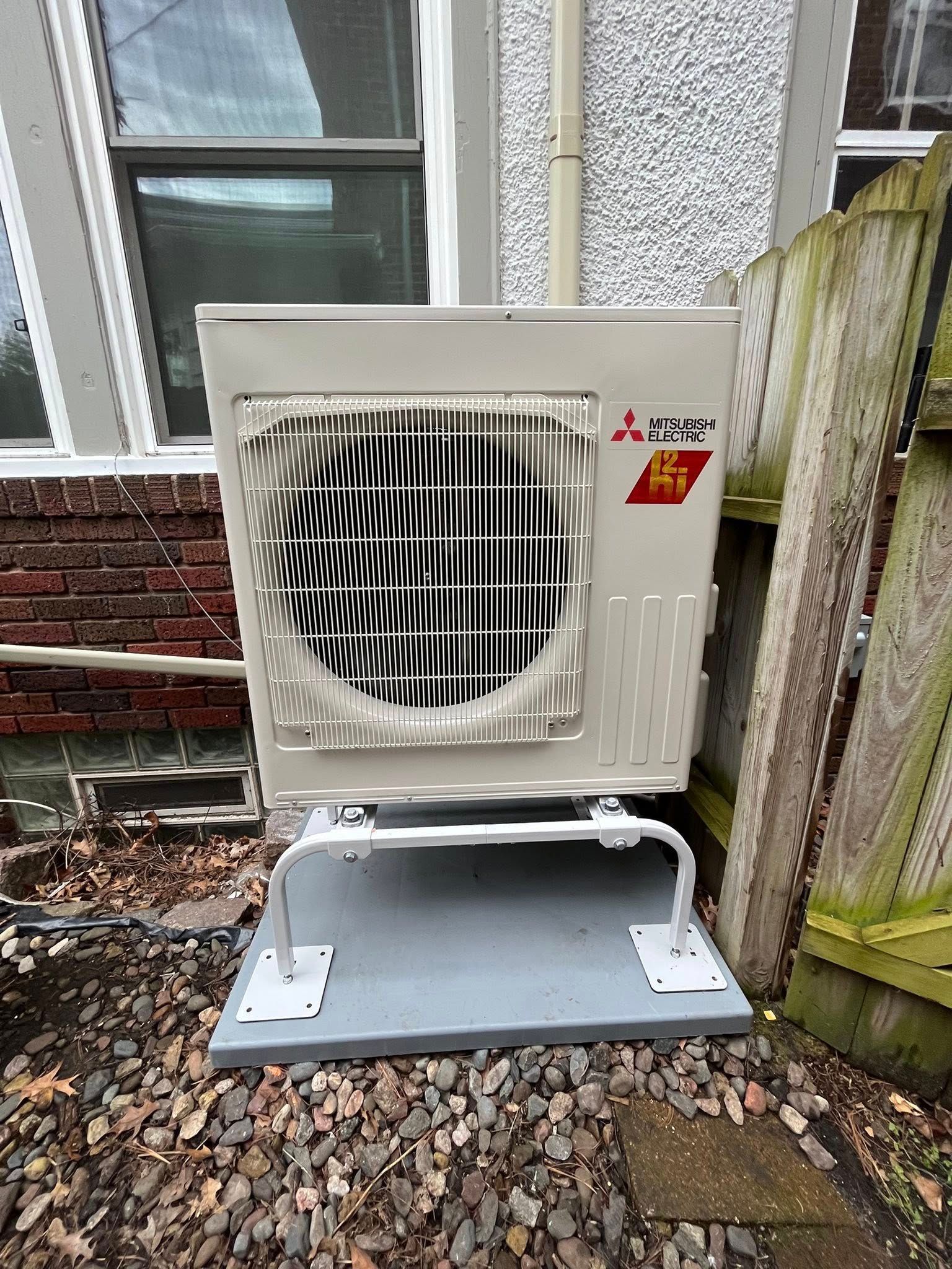 Mitsubishi outdoor HVAC unit, white, on a gray base, next to a wooden fence and brick wall.