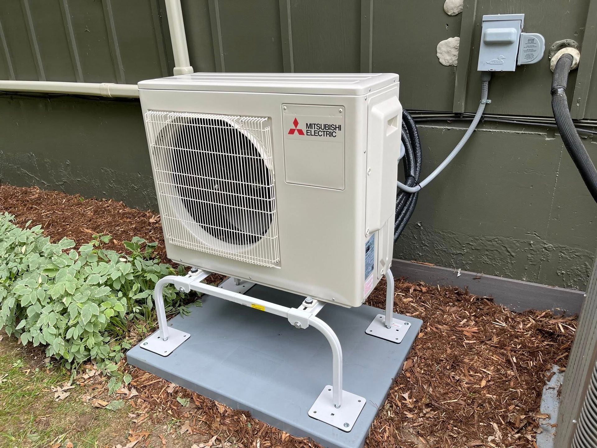 Mitsubishi heat pump unit, on a concrete pad, with supporting metal frame, near a building.