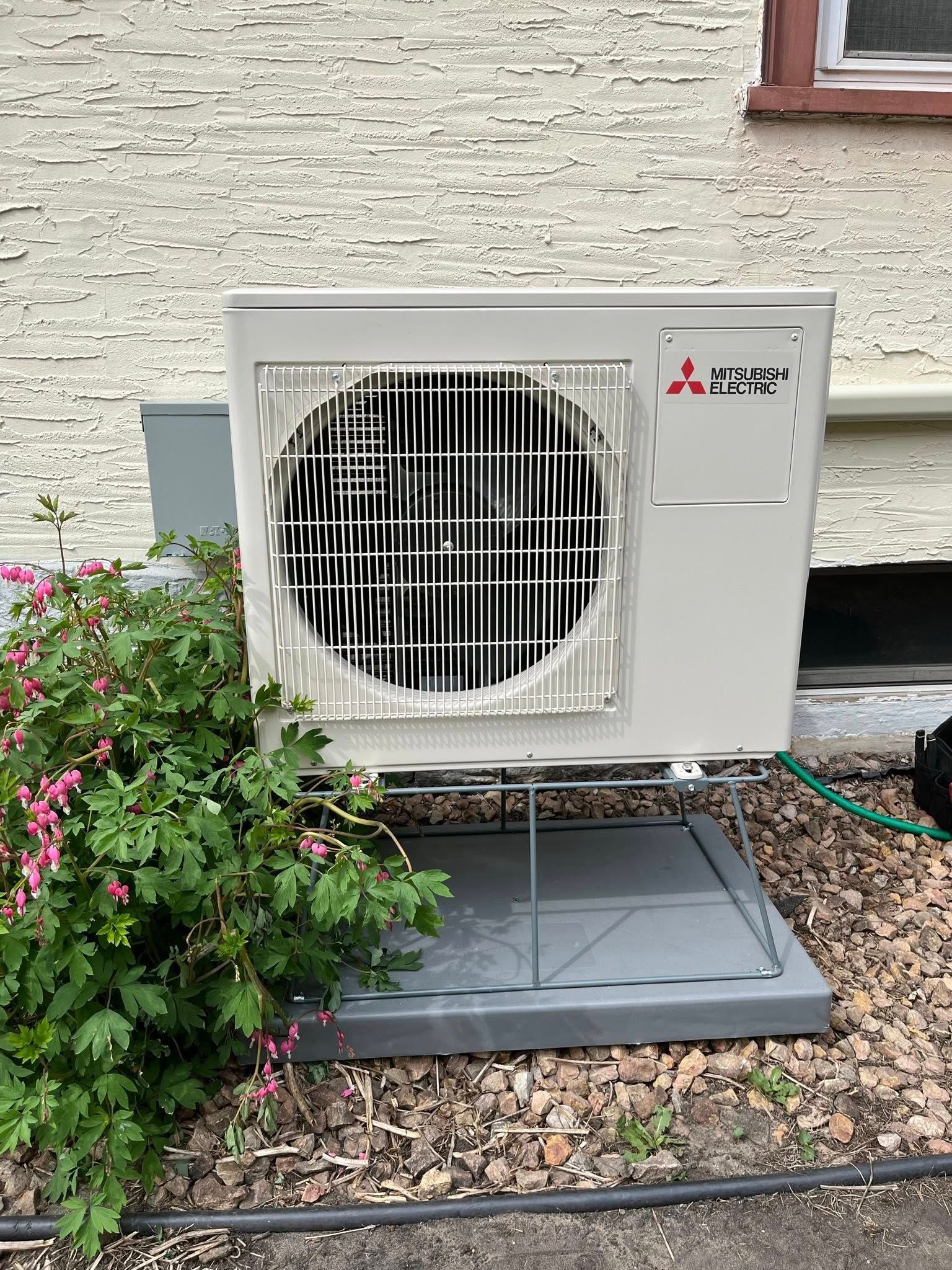 Mitsubishi heat pump unit mounted near a beige wall and bushes.