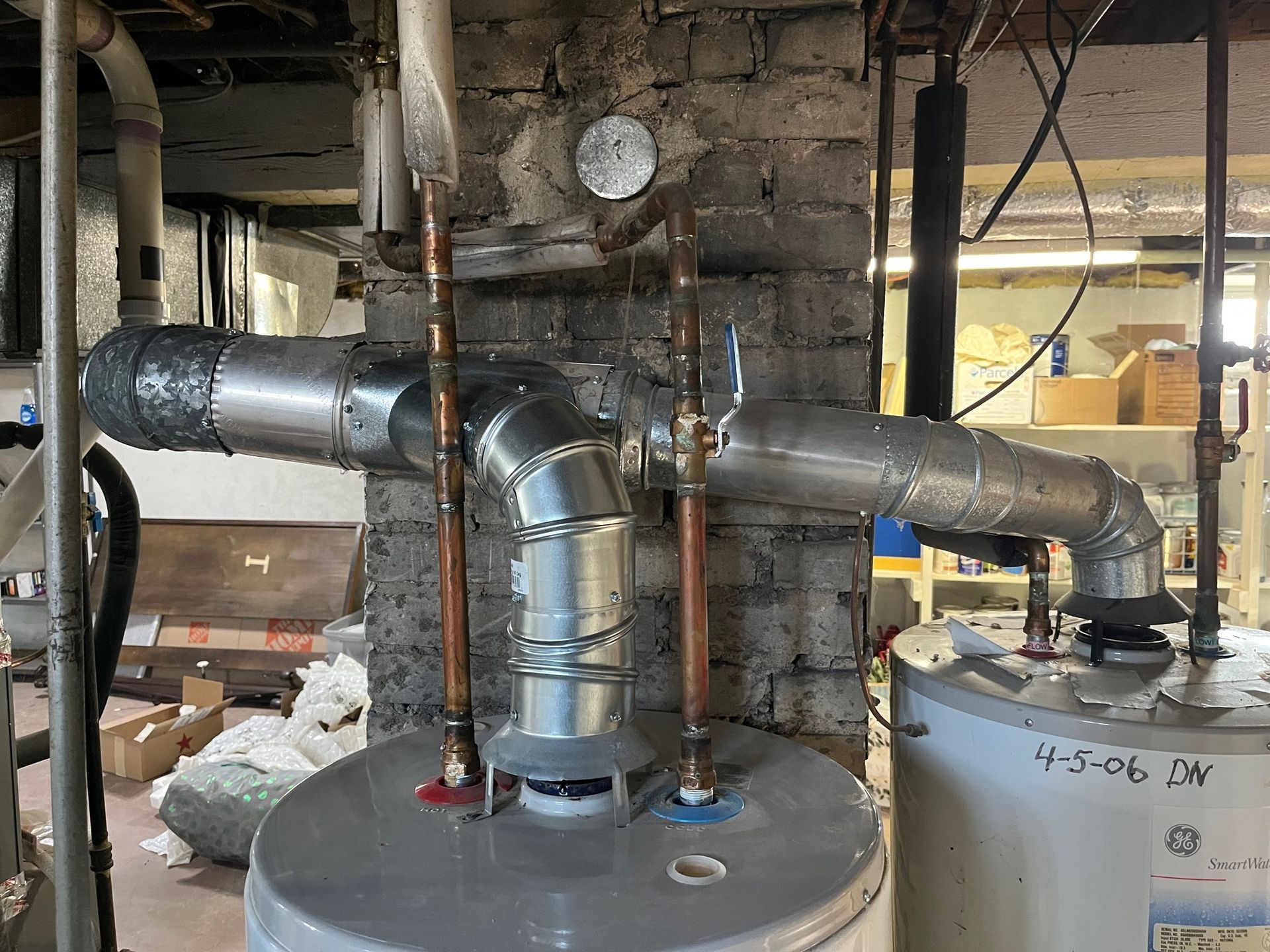 Two water heaters with copper pipes, insulated vents, and pipes against a brick wall in a basement.