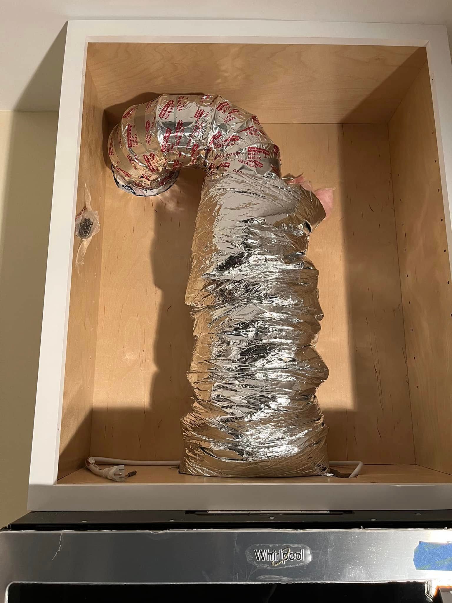 Accordion-like metal vent tubing inside a cabinet, connected to a kitchen appliance, reflecting light.