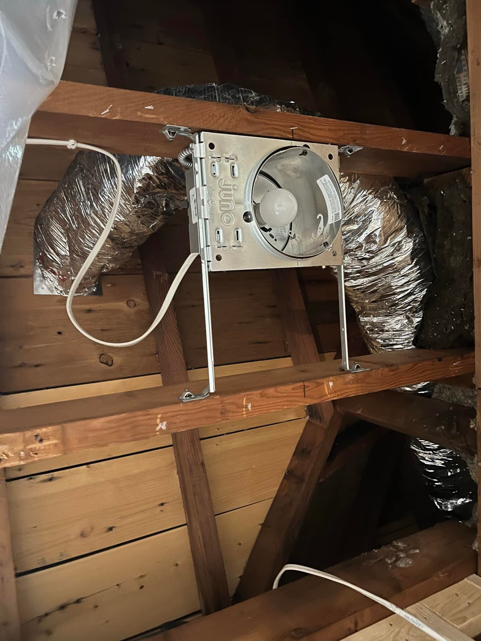 Attic view of an installed exhaust fan between rafters with flexible ducting and electrical wiring.