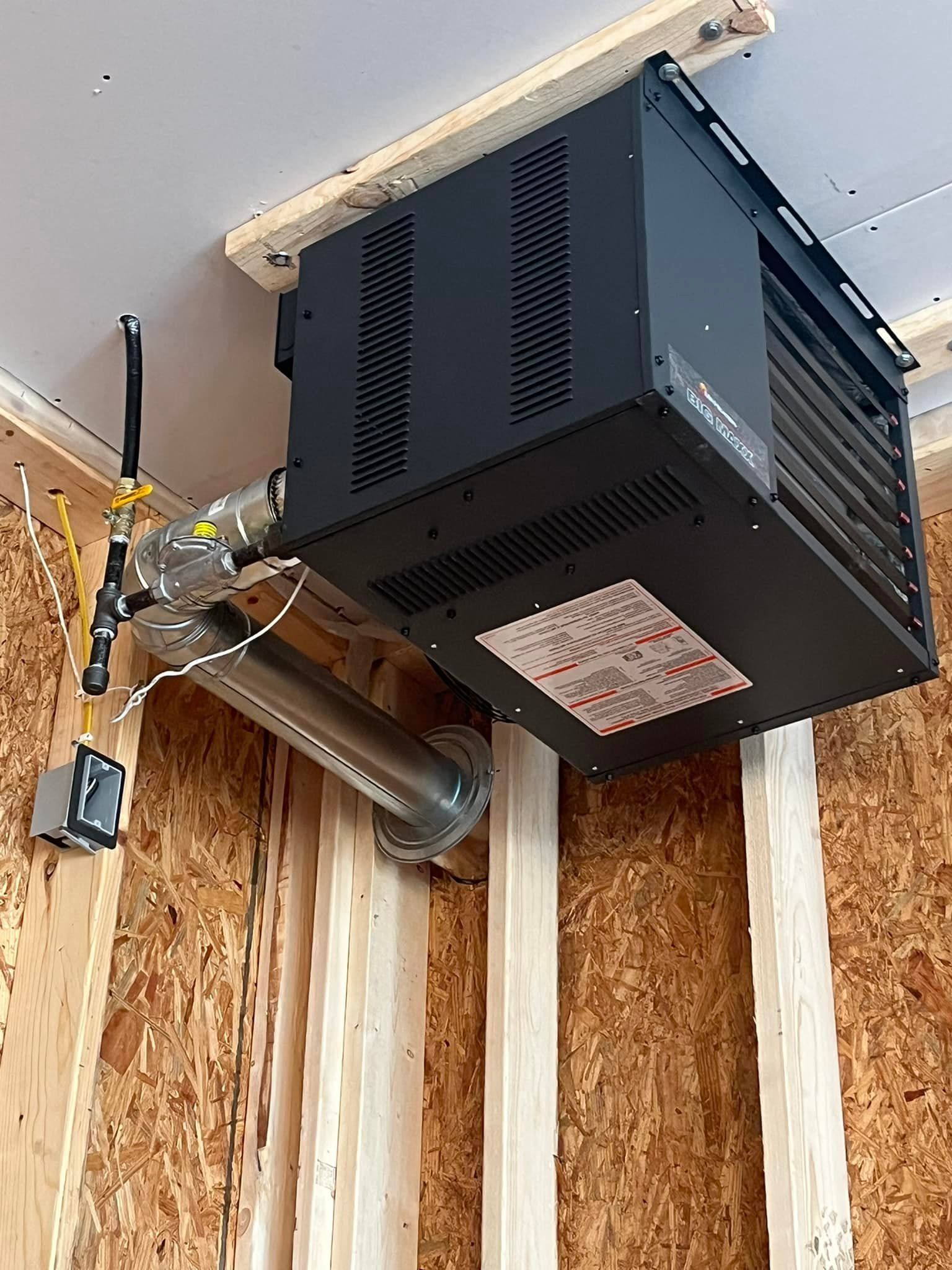 Ceiling-mounted black garage heater with metal exhaust pipe, attached to wooden beams and wall.