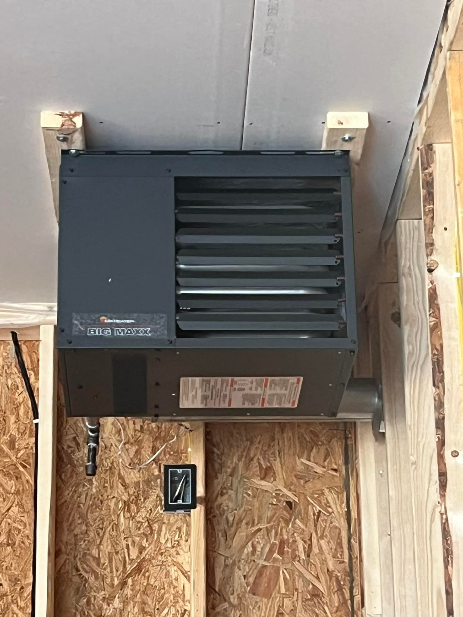 Ceiling-mounted gray garage heater installed between wooden beams and drywall. Electrical panel visible on the wall below.