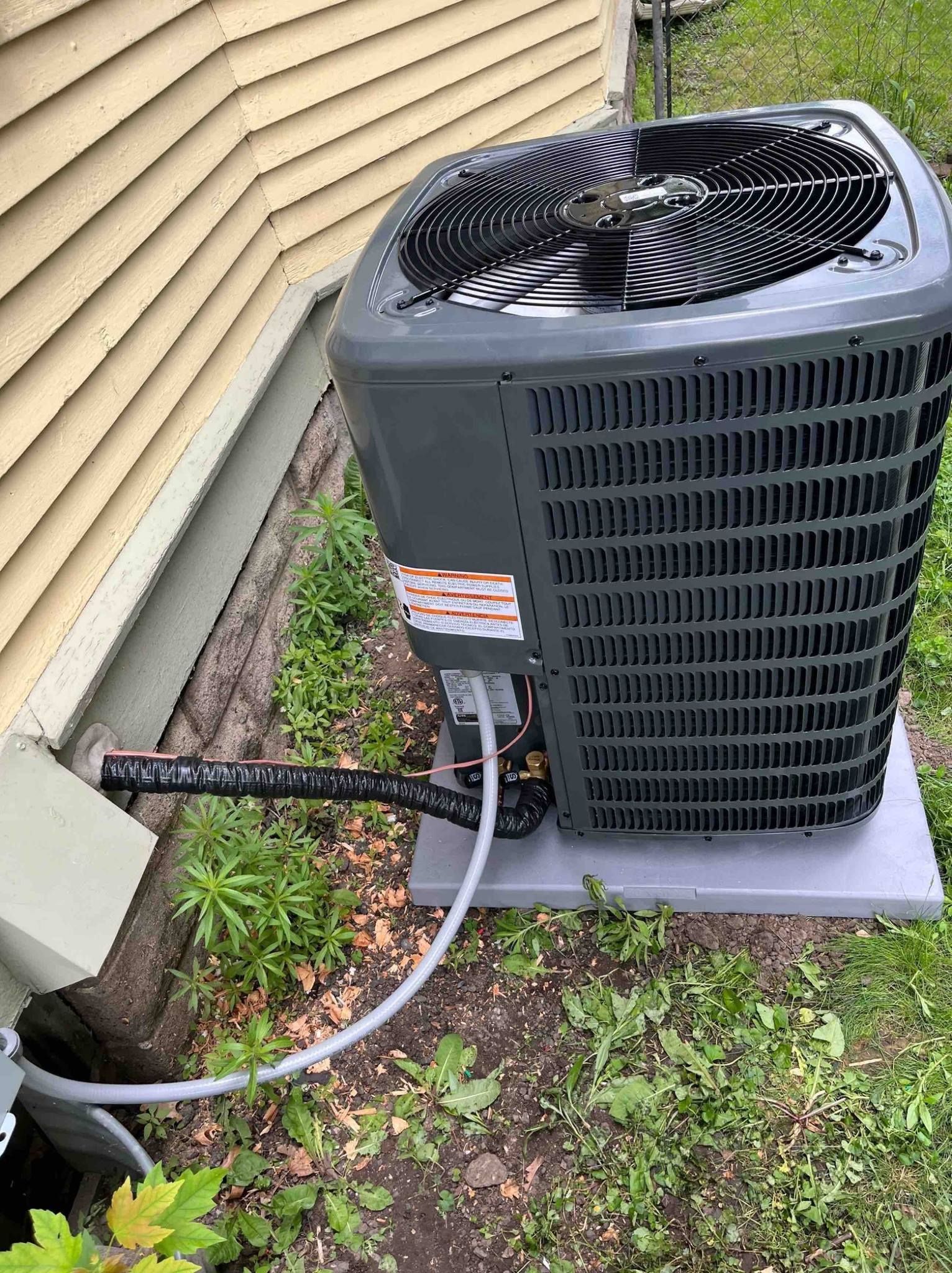 AC unit next to house, with black and gray components, surrounded by grass and plants.
