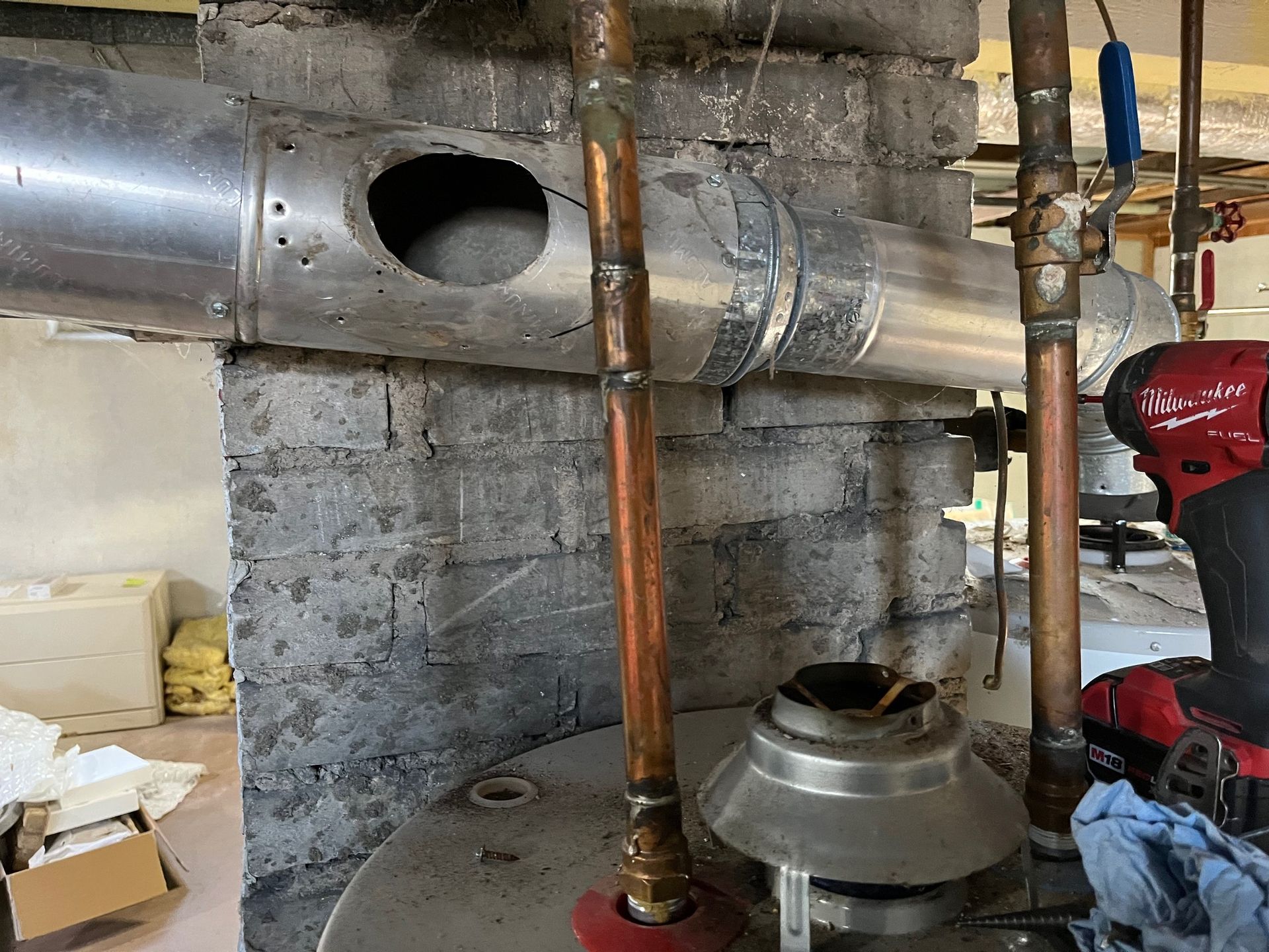 Water heater with copper pipes, a damaged vent, and brick wall in a basement.