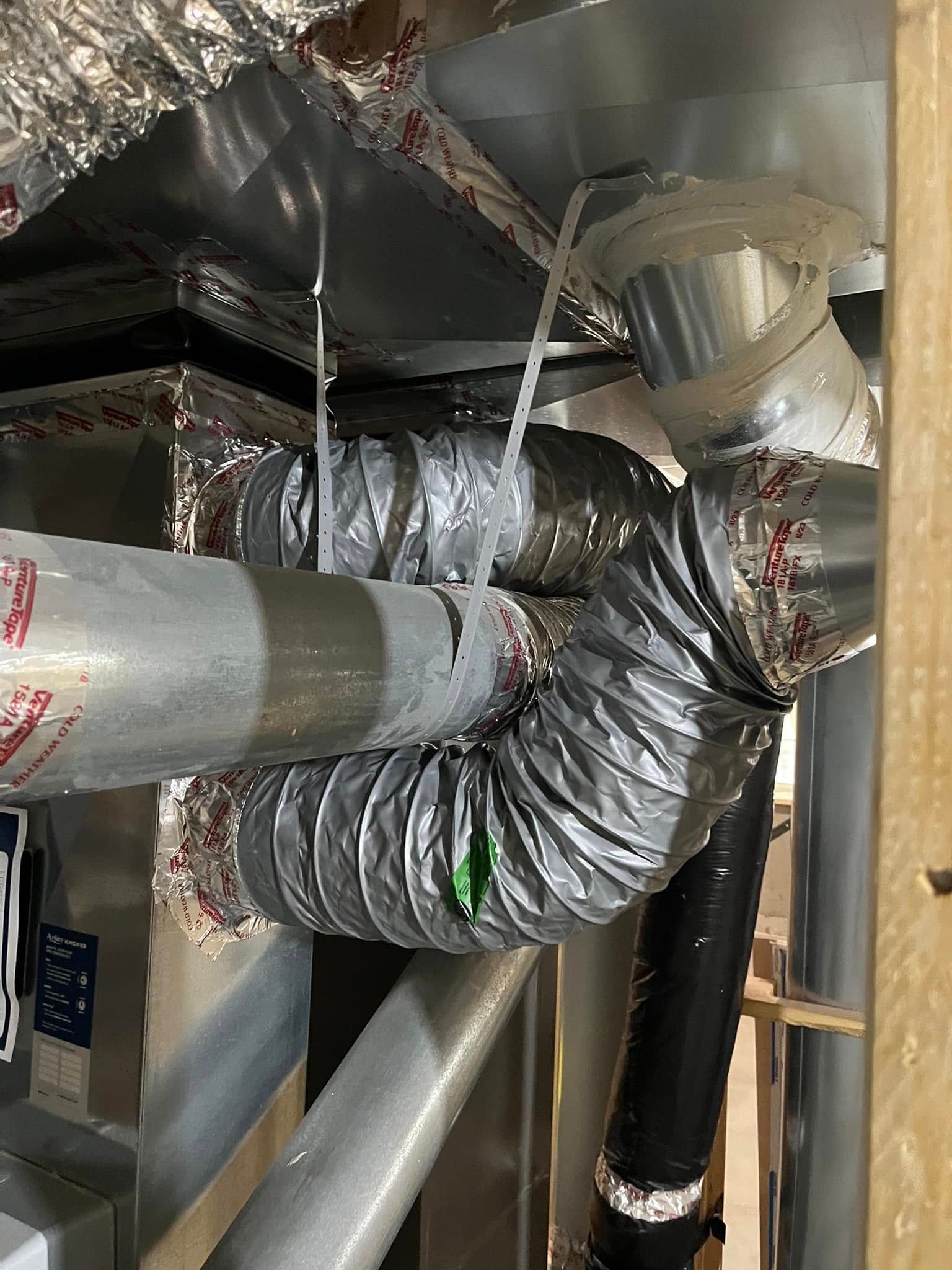 Flexible ductwork in a crawl space, connected with zip ties to rigid duct, silver and black pipes.