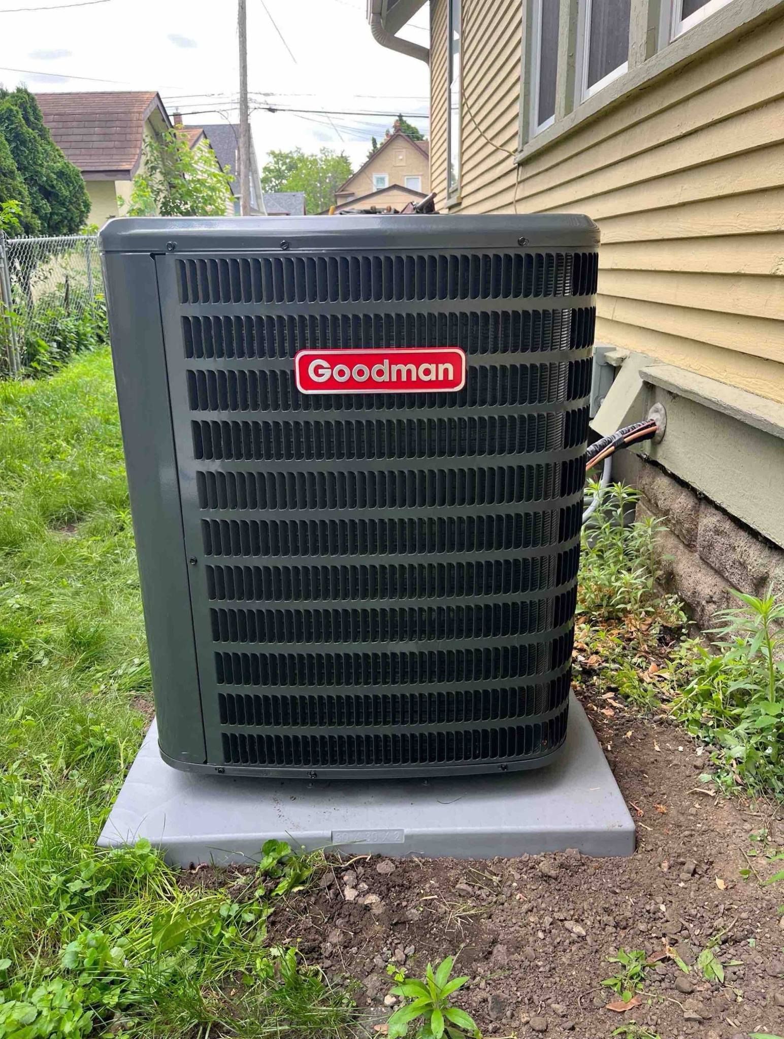 Goodman air conditioning unit on a concrete pad beside a yellow house with overgrown weeds.