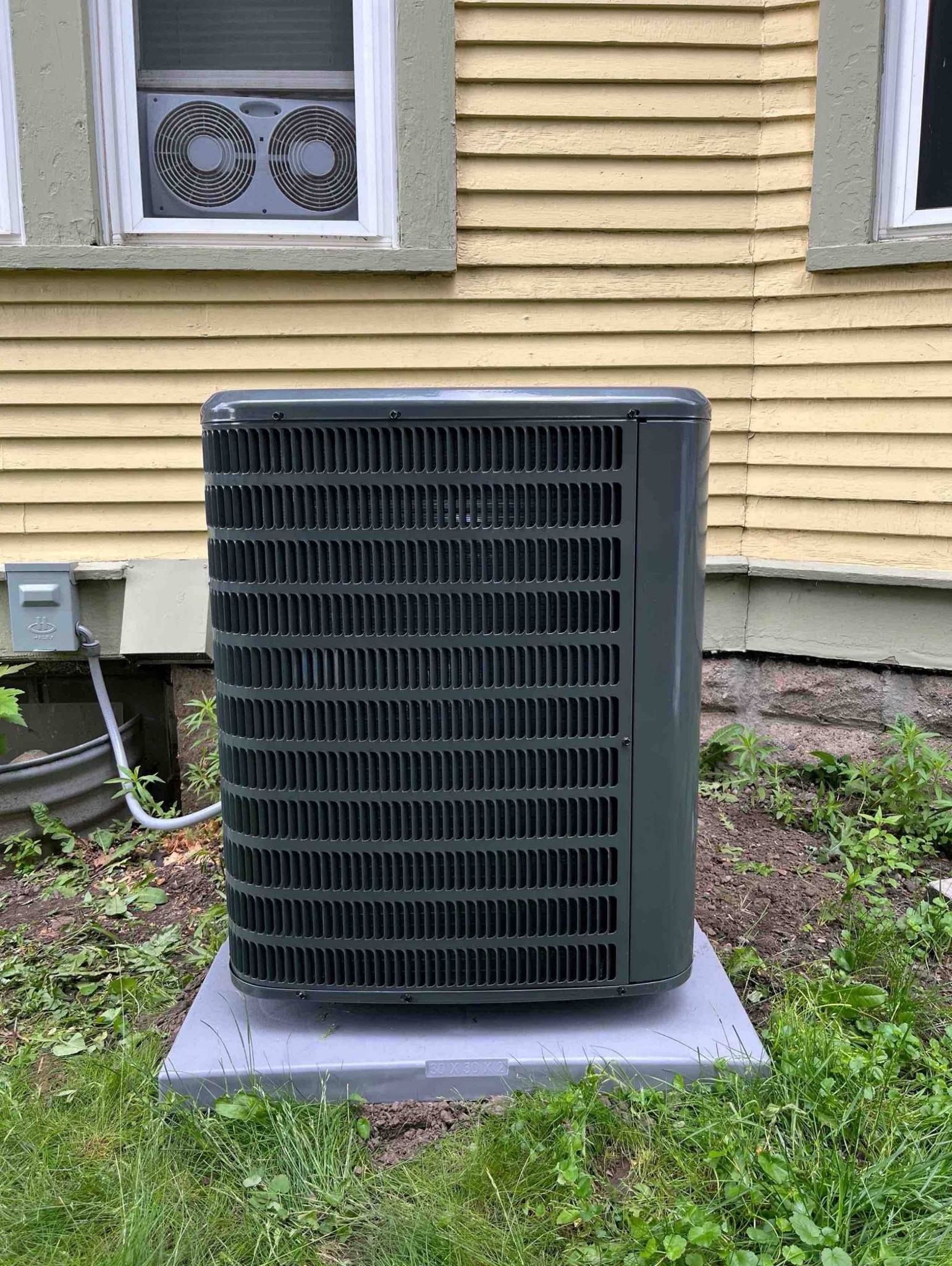 Air conditioning unit outside a yellow house, on a gray platform, surrounded by grass.