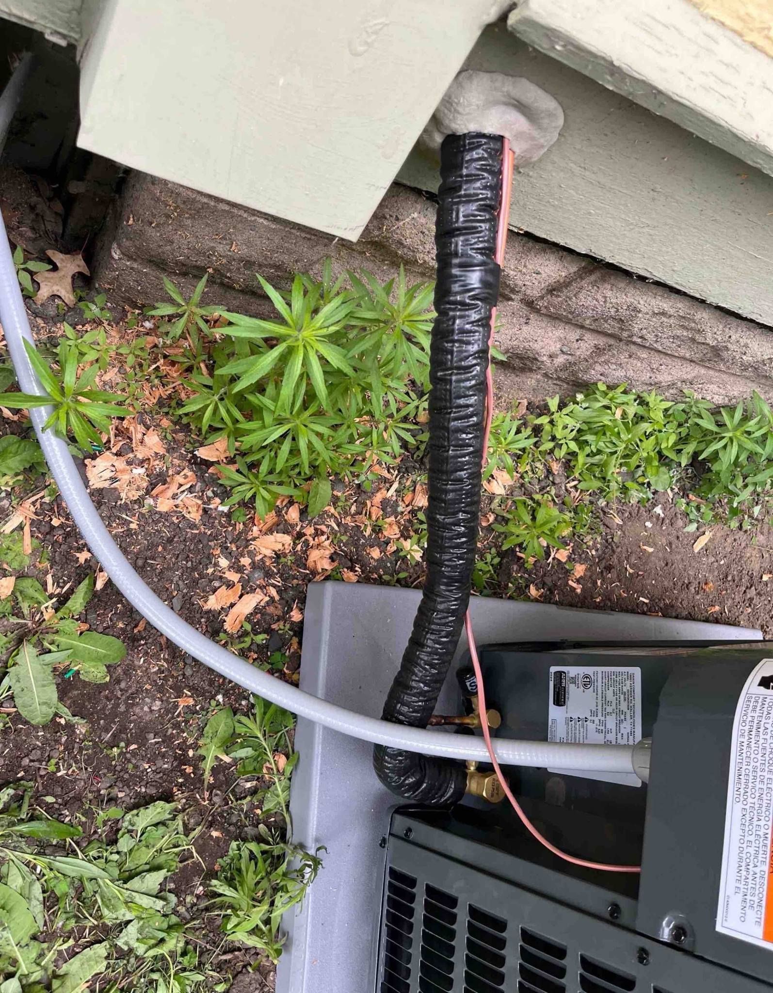 Black HVAC ducting and white tube near a building, with ground vegetation.