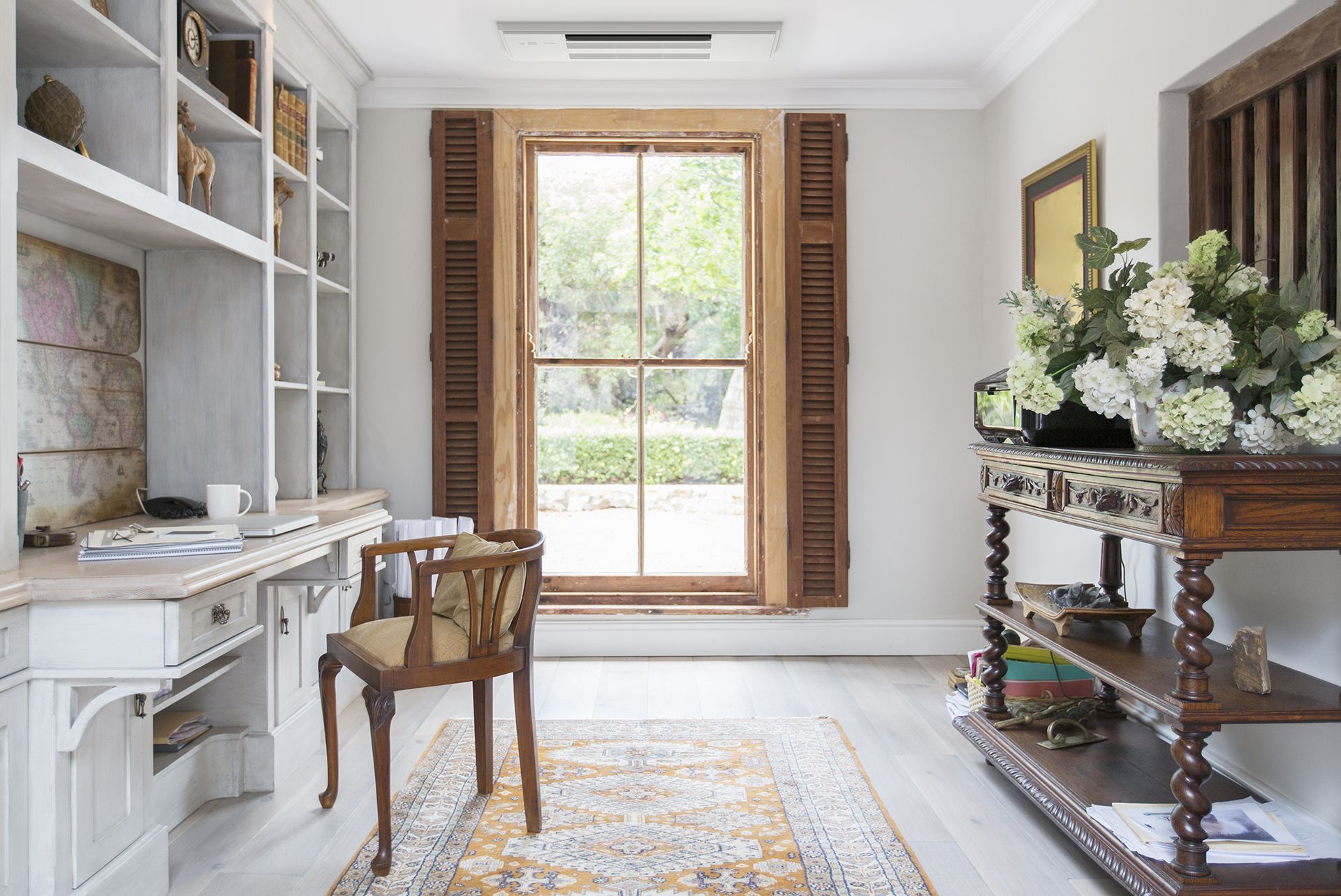 Desk with shelves, window with shutters, chair, and antique table in a home office.