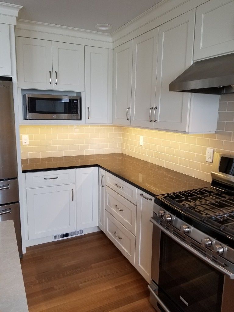 Modern white kitchen with dark countertops, wood floor, stainless appliances, and warm under-cabinet lighting
