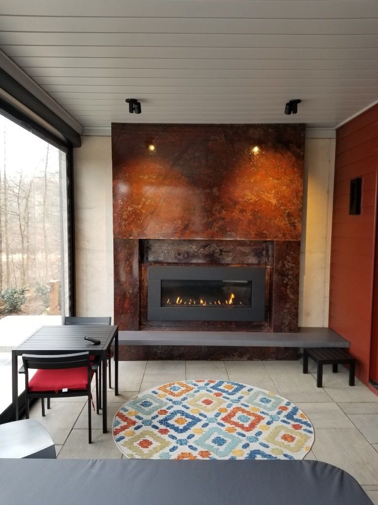 Covered porch with a copper-clad fireplace, tables, benches, and a colorful round rug.