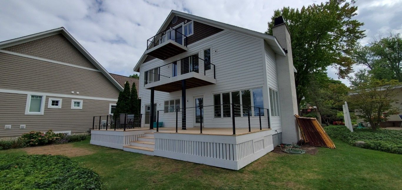 White multi-story house with front porch, black railing, and lawn beside a gray neighboring home