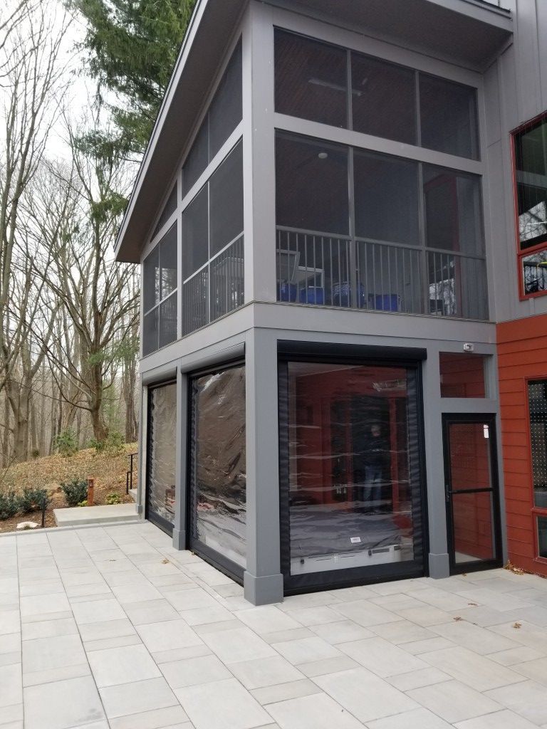 Modern two-story glass and gray building with a screened balcony and paved patio in a wooded area