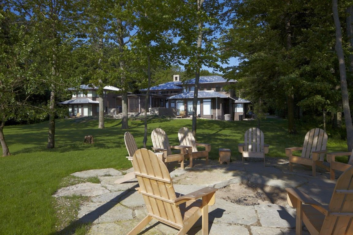 Outdoor patio with Adirondack chairs and a fire pit in front of a house, surrounded by trees and grass