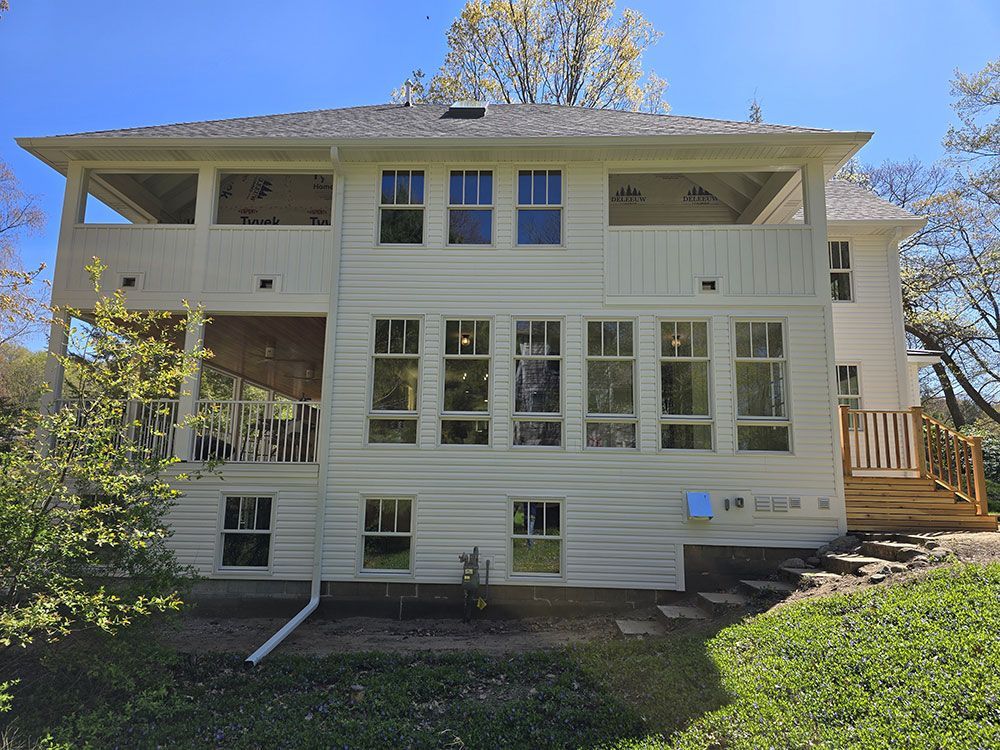 White two-story house with multiple windows, porch, and wooden deck. Sunlight, green grass, and trees.