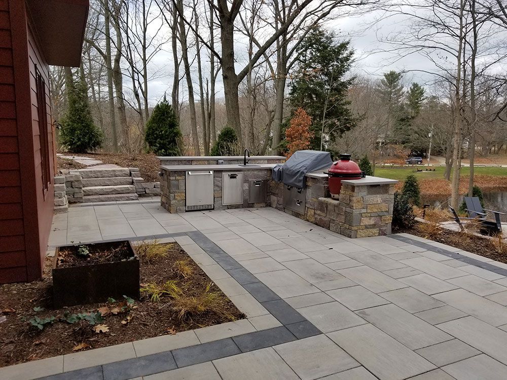 Outdoor kitchen on a stone patio with grill, sink, and wooded view.