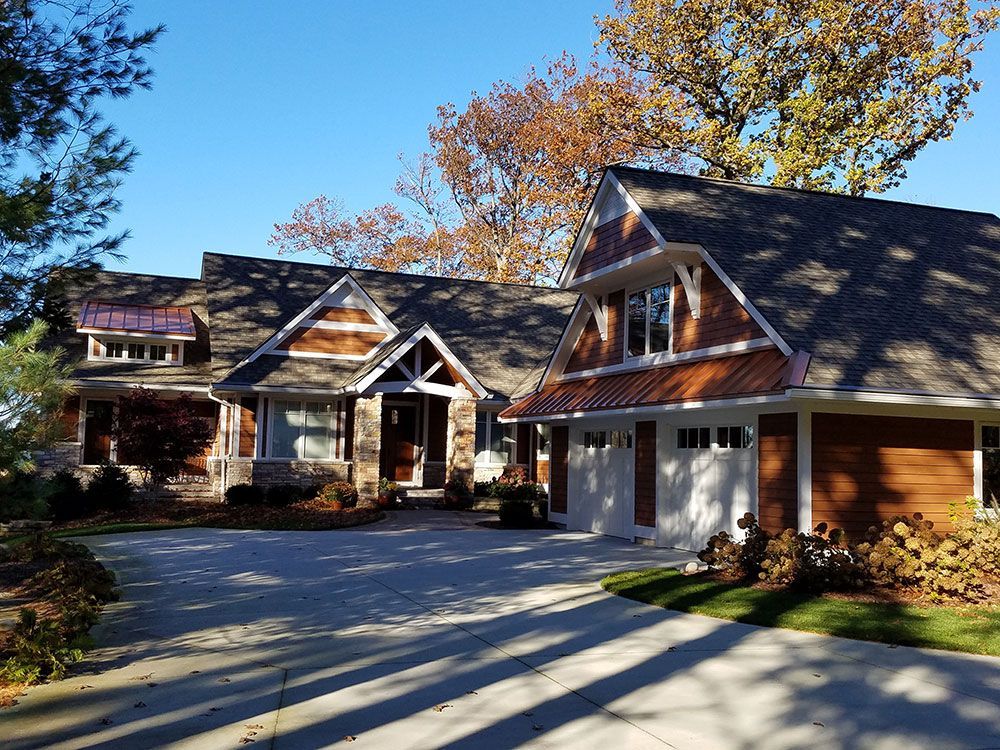 Brown-sided home with a white garage, stone accents, and a paved driveway on a sunny day.