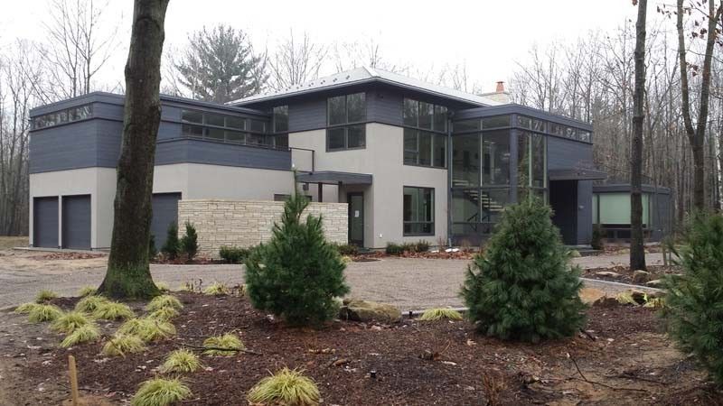 Modern two-story house with large windows, gray siding, and landscaped front yard in a wooded setting