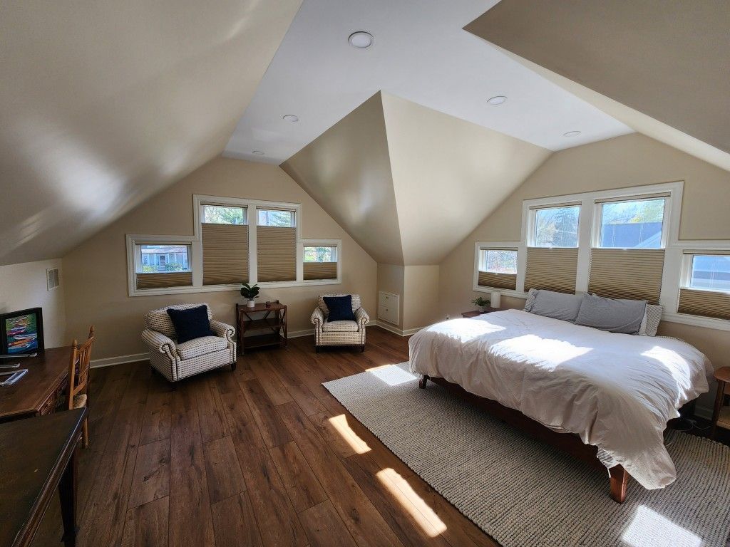 Spacious attic bedroom with sloped white ceilings, hardwood floors, and a bed by bright windows.