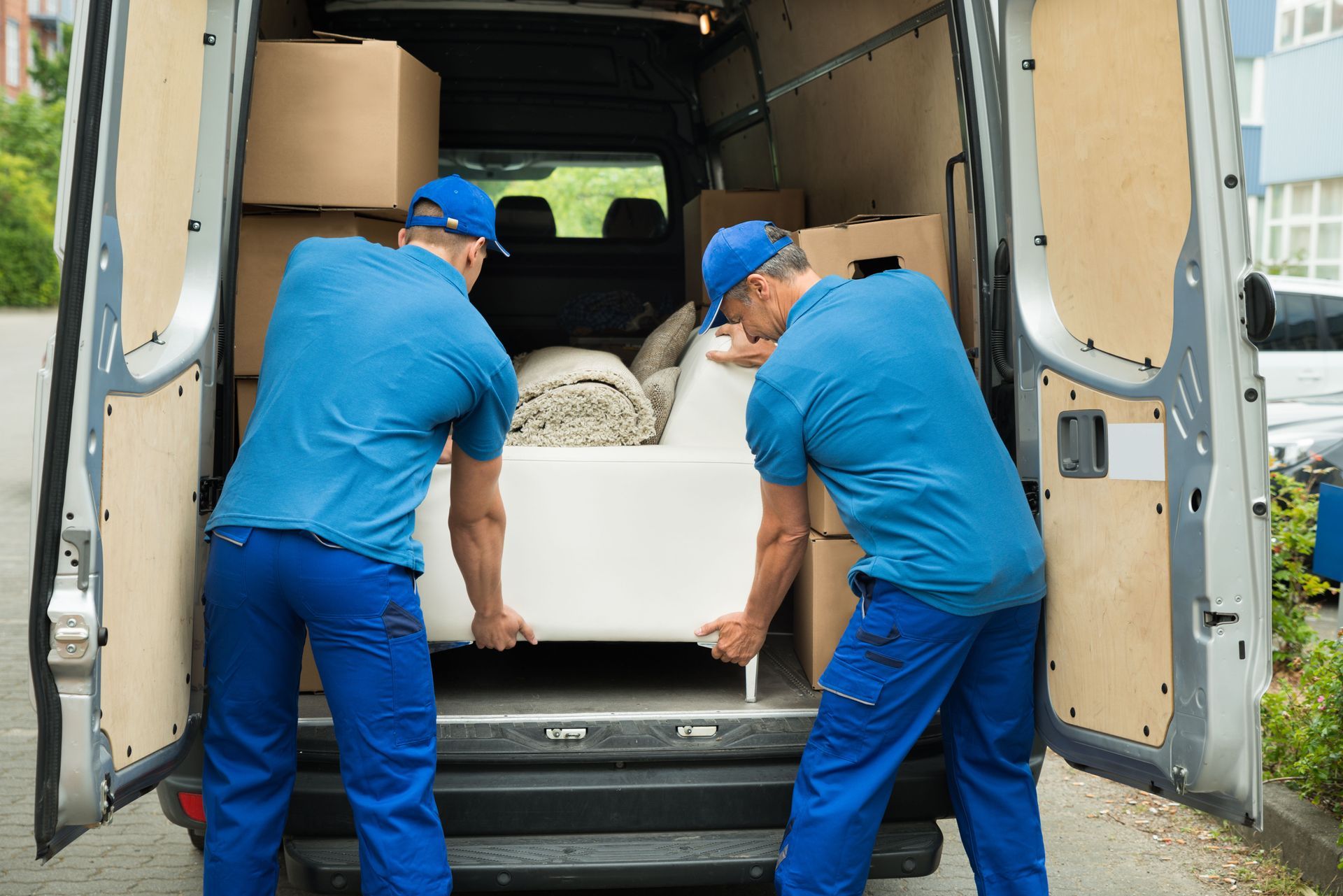 Two movers in blue uniforms loading a white couch into a van packed with boxes.