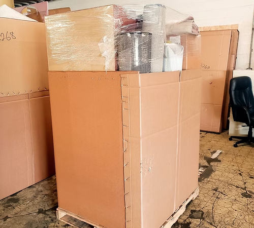 Large cardboard boxes stacked in a warehouse, with some boxes on a pallet, wrapped in plastic.
