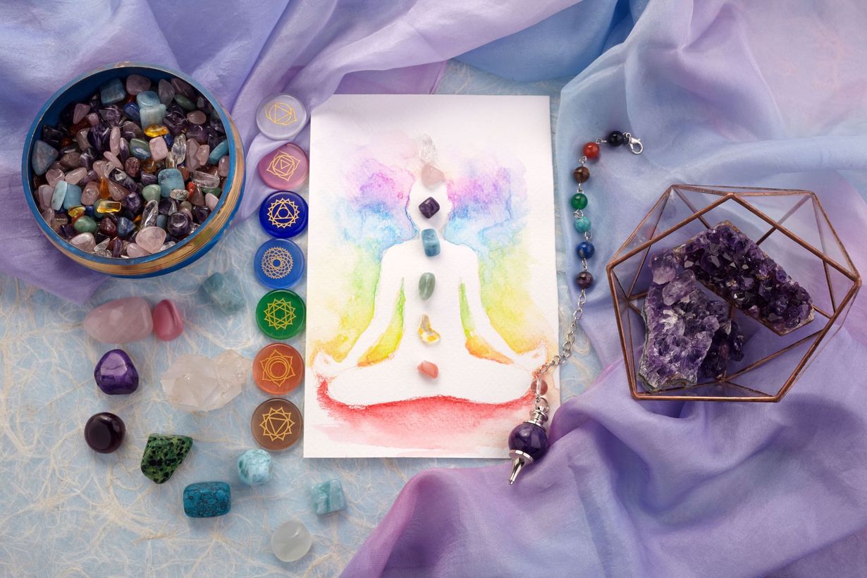 Arrangement of crystals, chakra chart, and pendulum on draped fabric, in shades of purple and pastel colors.