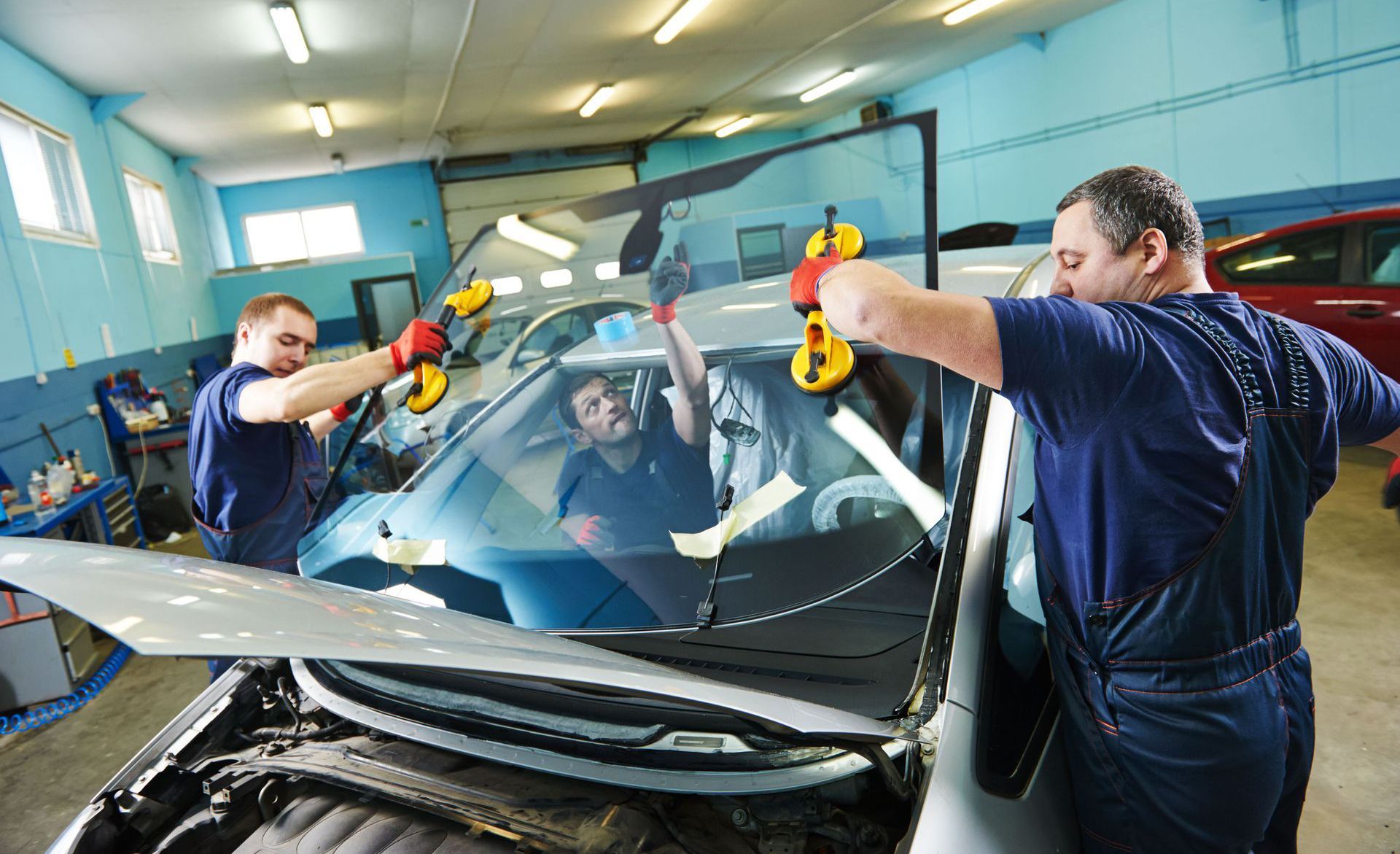 Three mechanics installing a windshield in a garage, using suction cups to position the glass.