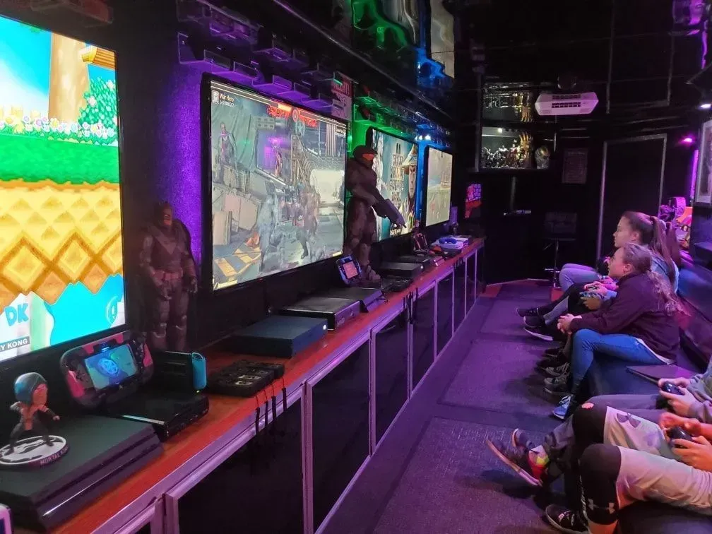 Inside a gaming trailer: Several screens showing video games; people sit on benches, controllers in hand. Purple and green lighting.