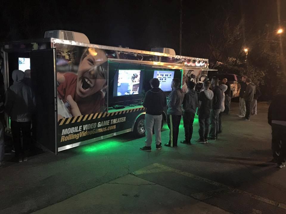 Mobile video game truck at night, with a line of people waiting to play. Green lights illuminate the underside.