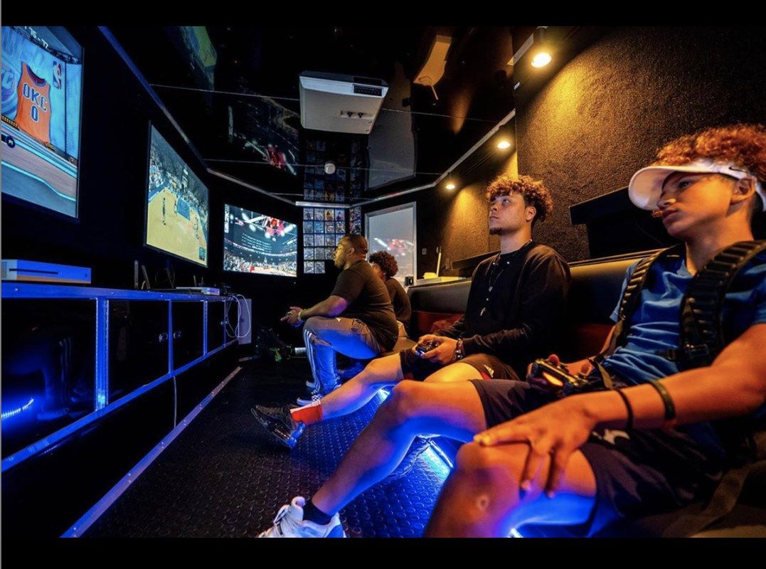 Three people playing video games inside a gaming truck with multiple screens and blue lighting.