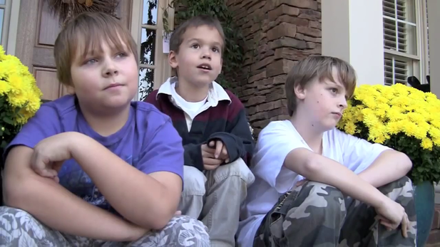 Three boys seated on steps, looking off-camera. Two have arms crossed. Yellow flowers flank them.