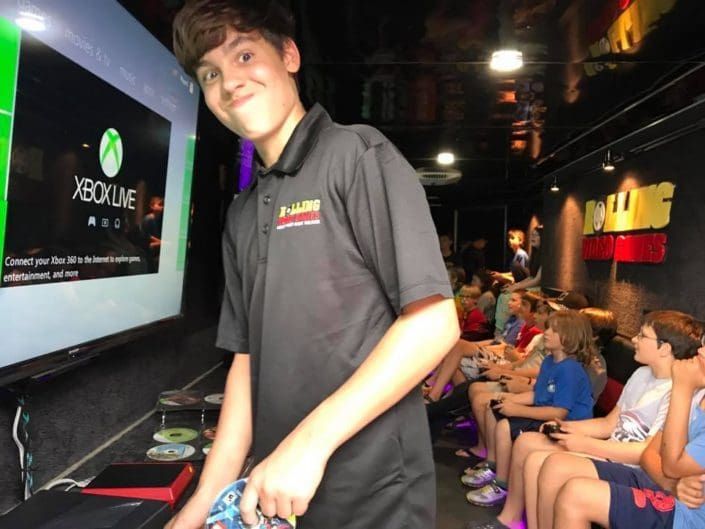 Teen holding a controller smiles in a gaming truck with other kids watching screens.