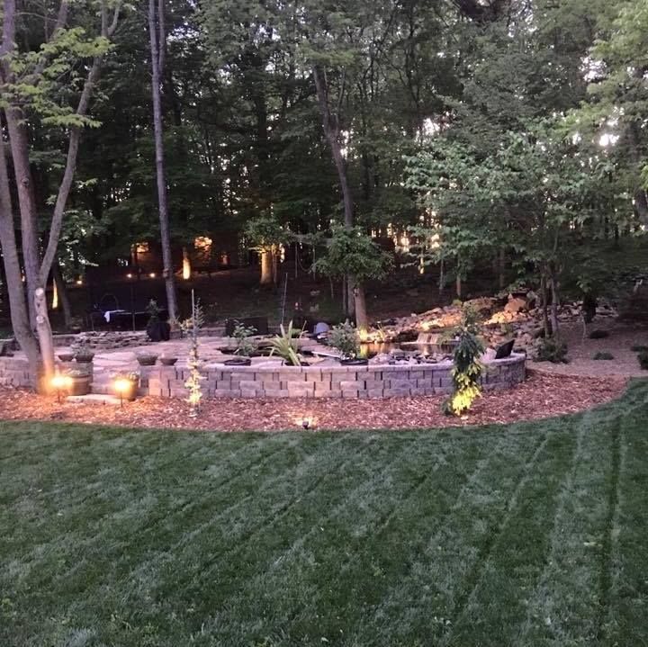 Lit backyard patio with stone retaining wall, warm landscape lights, and wooded trees at dusk