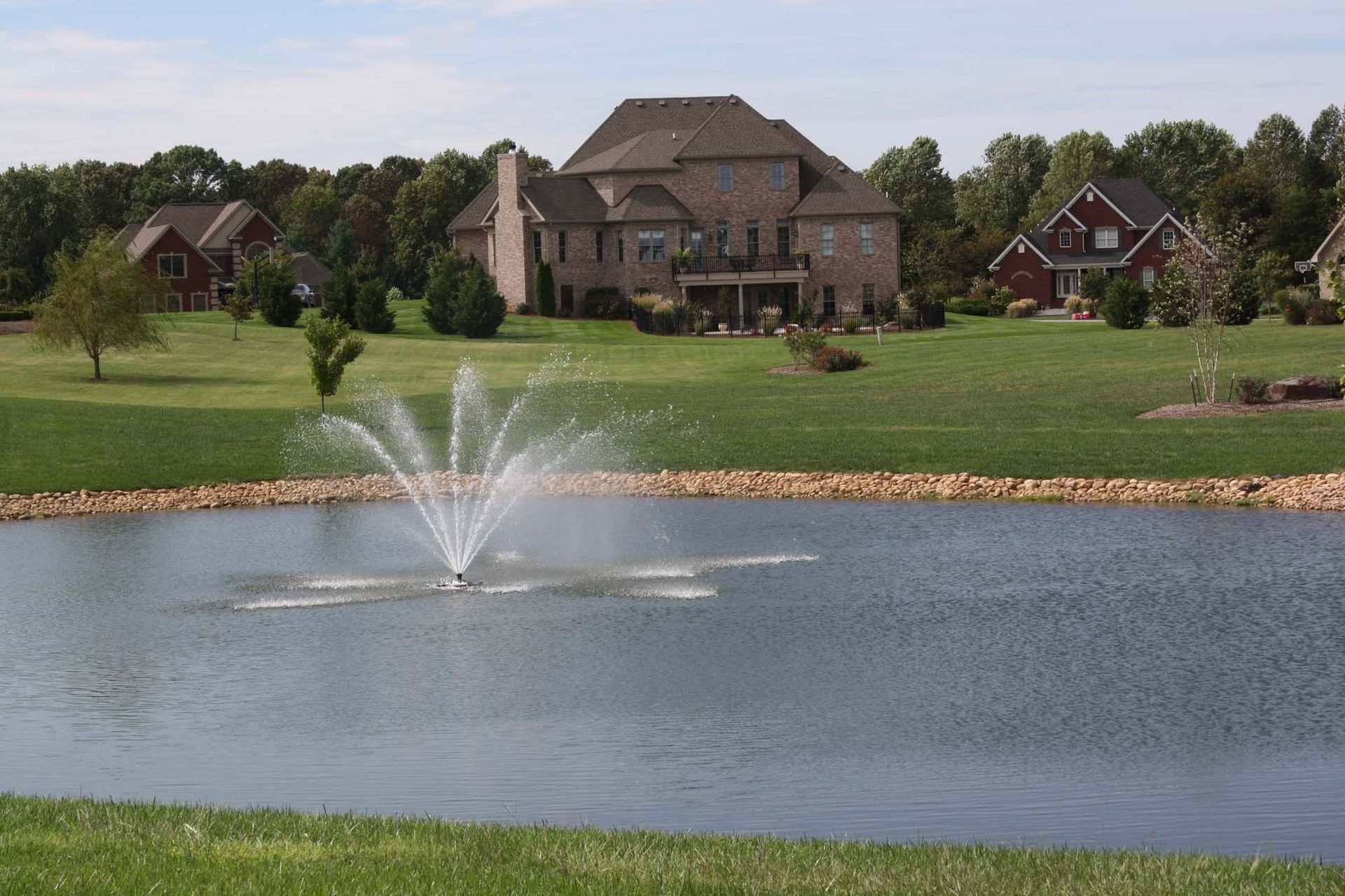 Large house across a pond with a water fountain and green lawn under a clear sky
