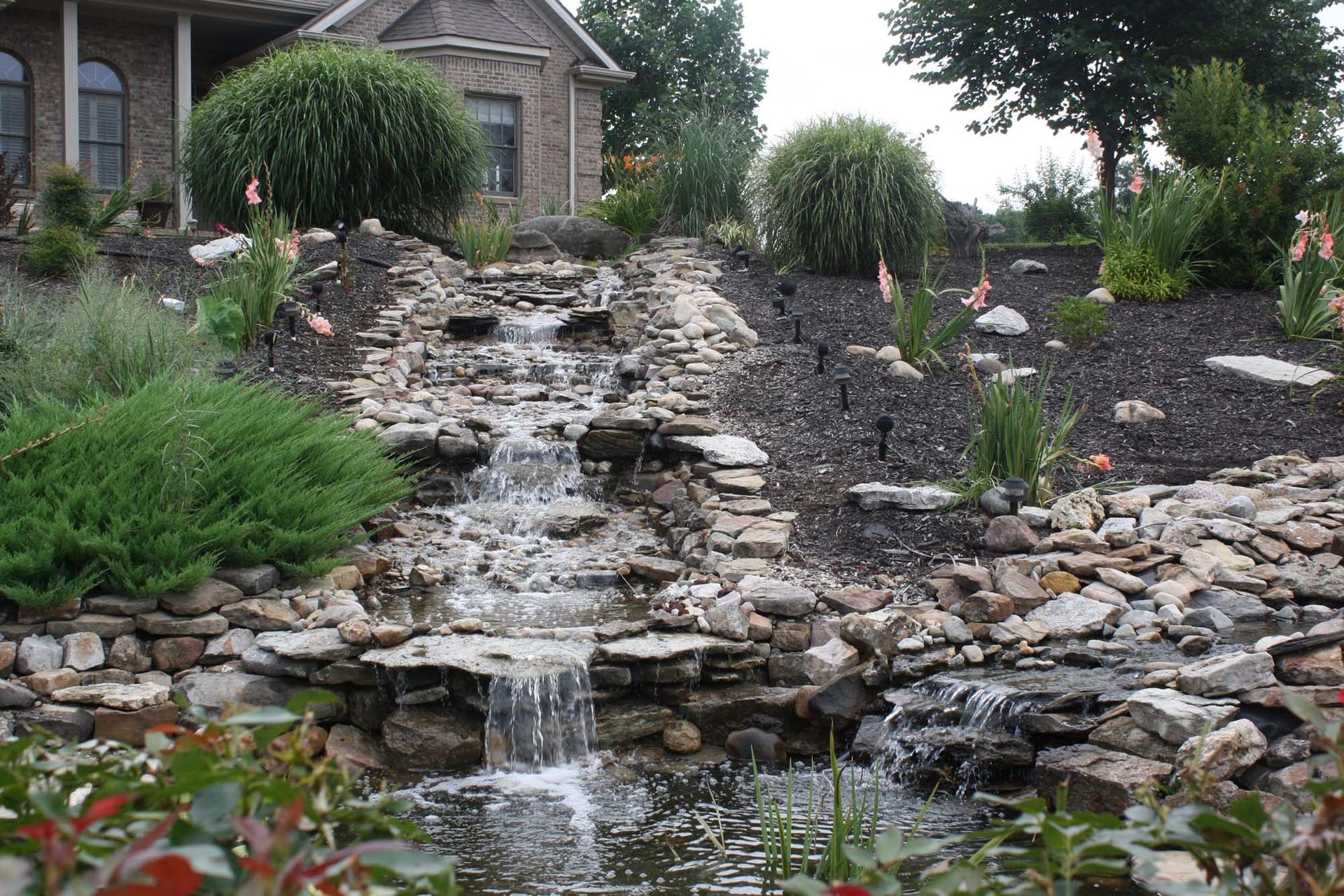 Rock garden waterfall cascading over stacked stones beside a house and landscaped shrubs
