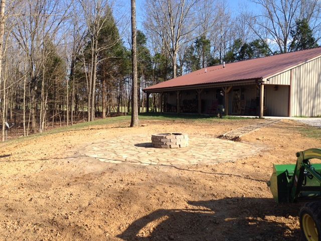 Dirt yard beside a metal-roofed building, with trees in the background and a fire pit in the center.