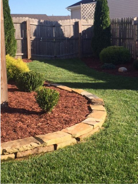 Backyard garden with curved stone border, mulch bed, shrubs, grass lawn, and wooden fence under sunlight