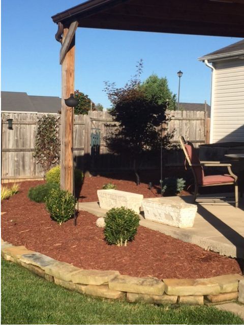 Backyard garden bed with mulch, shrubs, stone edging, and a wooden pergola beside a patio and fence