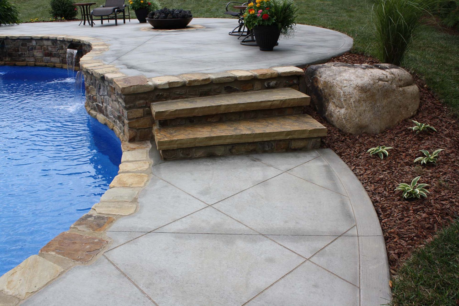 Curved concrete path beside a blue pool, leading to stone steps and a patio with potted flowers.