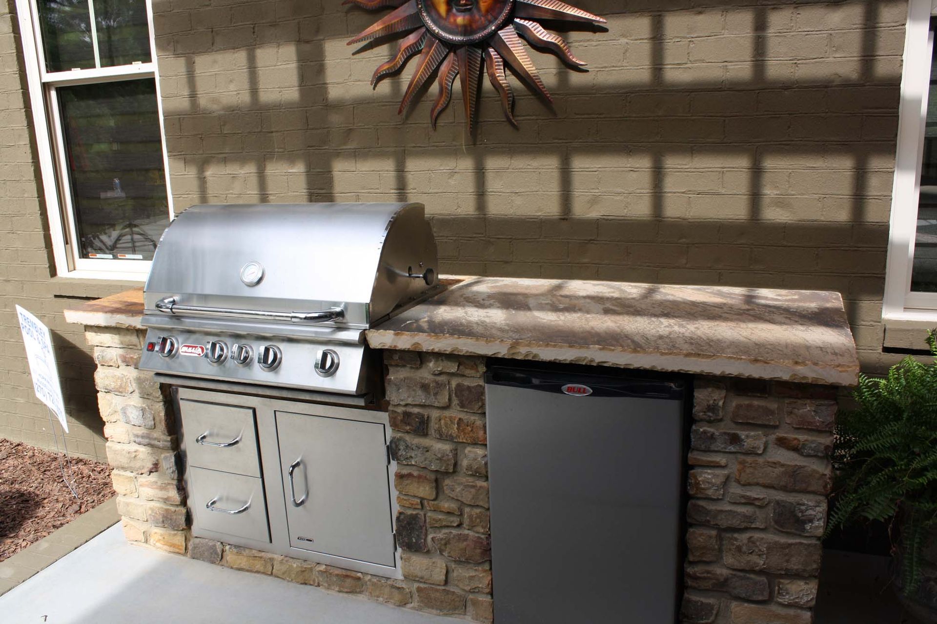 Patio with stainless steel grill, black trash bin, and stone wall beneath a window and sun decoration